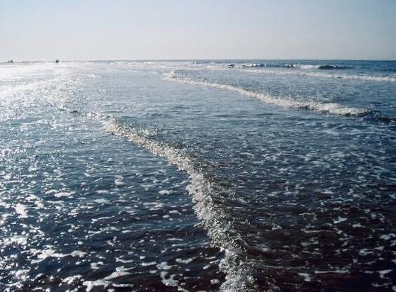 Seichte Wellen rollen flach über den Strand und scheinen die Füße der Fotografin zu umspülen. Es ist Flut. Lediglich hinten links im Bild ist ein bisschen glänzend nasser Sand zu sehen über den in der Ferne ein Pärchen entlang schlendert. Ihre dunklen Silhouetten setzen sich gegen den hellen Sand und den blauen Himmel ab.