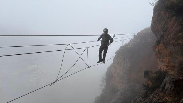 A person walks a high line wire bridge over a chasm surrounded by cloud and mist