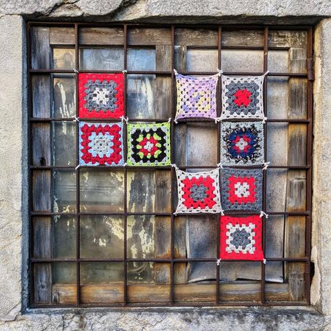 A weathered window with a rusty metal grid has nine colourful crocheted granny squares tied to it with white yarn. The squares are arranged in a 3x3 grid. From left to right, top row features a red and grey square, a pastel purple and cream square, and a grey and red square. The middle row displays a red and light blue square, a black, green, and yellow square, and a blue, red, and white square. The bottom row shows a red and grey square, a red and white square, and a red and grey square. The window itself is aged, with peeling paint and grime on the glass panes. The surrounding stone frame is also worn, suggesting the window is part of an old building.