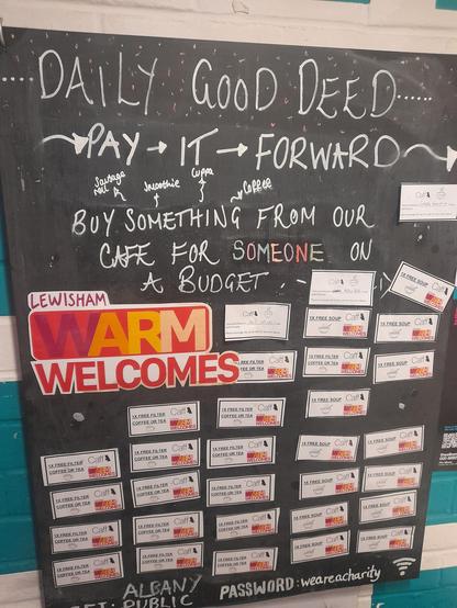 a chalkboard with “daily good deed. Pay it forward. Buy something from our cafe for someone on a budget” written on it. A large sticker for Lewisham Warm Welcomes is stuck to it and the rest of the board is filled with smaller stickers of pay it forward items.