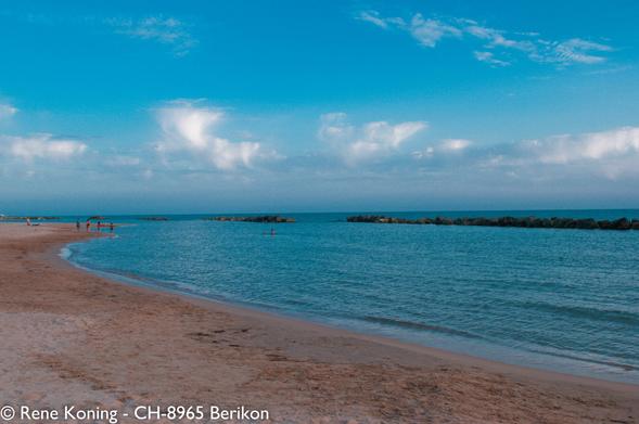The image depicts a serene beach scene with a clear blue sky and gentle waves lapping at the shore. The sandy beach curves gently, with a few people visible in the distance, some walking along the water's edge and others swimming. The water is a calm turquoise, with a breakwater of large rocks visible in the midground, creating a natural barrier. The sky is mostly clear with a few scattered white clouds, adding to the tranquil atmosphere. The overall color palette is dominated by blues and sandy tones, contributing to the peaceful ambiance of the scene.