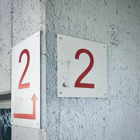auf dem bild sind zwei schilder mit der roten zahl „2“ zu sehen, die an einer grauen, rauen ecke eines hauses befestigt sind. das schild links zeigt zusätzlich einen pfeil, der nach oben rechts abknickt. im hintergrund, rechts oben an der wand, ist außerdem eine erhabene zahl „2“ direkt auf der wandoberfläche zu erkennen. also dreimal die zwei als hausnummer.