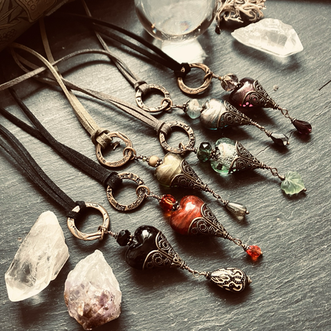 A moody photo of six pendants on rough slate surrounded by crystal points and a crystal ball. The pendants are elaborately armoured glass hearts with crystal glass beads crowning them like sacred hearts, layered with more antique brass filigree. They hang from faux suede cord