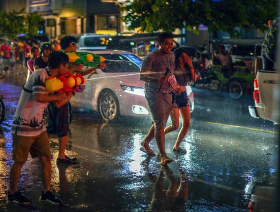 Photo shows a couple walking in a dark street lighten up by car headlights and street lights. It is wet and water splashing everywhere, by people throwing water from bucket and shooting from water guns. The couple walks hand in hand bending over and keeping up hands and arms to shield themselves from the worst of water splashes.
