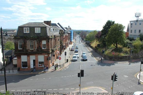 View along a road, from an elevated position near a crossroads. On the left are 3 storey brick buildings( 1920s in design), on the right a wooded plot around  4 storey modernist block