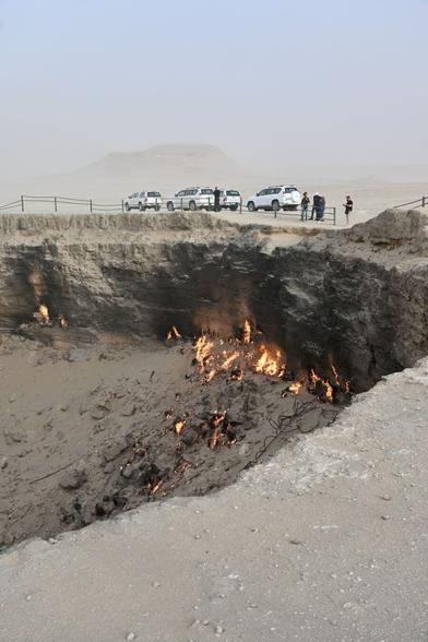 A crater in the desert with flames in it. At the rim there are some white 4WDs parked and people gazing down.