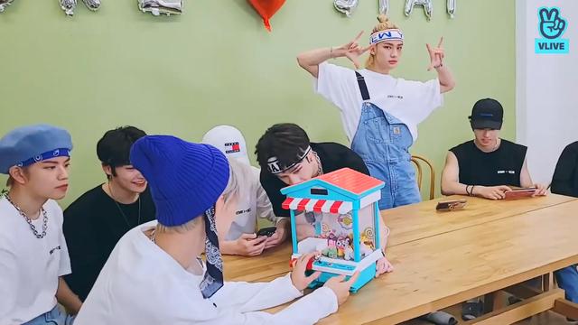 skz members sit around a table and play a table-top claw game. Hyunjin is standing up and dancing to the claw machine's tinkly music.