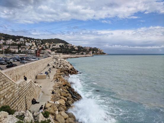 A wide, slightly elevated shot captures a coastal scene with a rocky breakwater extending into the sea. The sky above is partly cloudy, with patches of blue visible amidst the white and grey clouds. The sea is a muted turquoise-grey, with white foamy waves crashing against the large, irregularly shaped rocks that form the breakwater.
Along the top of the breakwater, a paved walkway runs parallel to the sea. Several people are visible along this walkway; some are standing and looking out at the water, while others are sitting on the stone edges. A low stone wall lines the inland side of the walkway.
Behind the walkway, a multi-tiered stone structure rises upwards, leading to a road where numerous parked cars are visible. Beyond the road, a hillside covered with buildings in various shades of beige, white, and terracotta stretches into the distance. The buildings appear densely packed, indicating an urban or residential area along the coast.