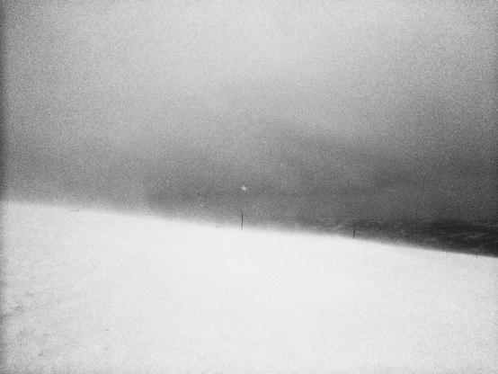 Snow-covered slope during a blizzard, with strong grain and low visibility. A few thin poles are faintly visible in the distance, marking the edge of the ski route. The horizon is obscured by dense snowfall and grey sky. No people or objects are clearly discernible.