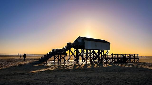 At sunset, a stilt house stands gracefully on the beach, casting long shadows across the sand. The warm light behind the roof forms a radiant silhouette, enveloping the scene in golden hues.