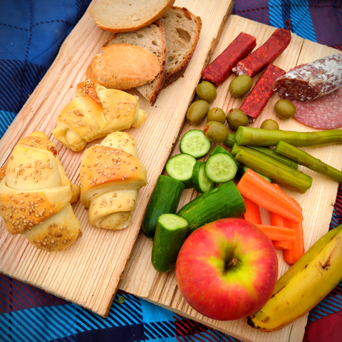 Auf Holzplatten ist ein Picknick mit Brot, Hörnchen, Apfel, Banane, Oliven, Wurst, Gurken, Karotten und Spargel angerichtet.