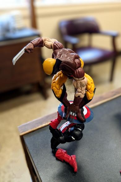 A Marvel Legends Wolverine in the yellow and brown suit balancing on US Agent's shield. US Agent appears to be struggling to hold up the weight