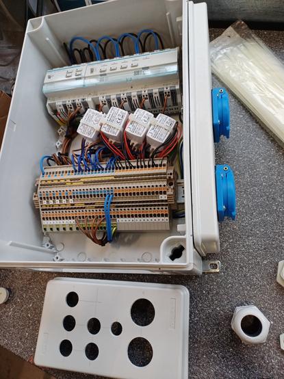 Wired plastic electrical enclosure containing a KNX setup.

At the top, there is a 4-channel switching actuator on the left and a 4-channel dimming actuator on the right, both from Hager. Below them are twelve small white power supplies. The terminal blocks are now wired and organized.

On the right side of the enclosure, two blue sockets with protective lids are mounted. A plastic bottom plate with multiple circular cutouts, intended for cable entries, is placed next to the enclosure on the workbench.