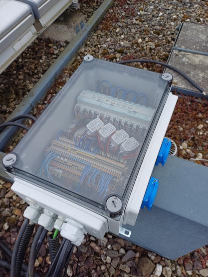 Electrical control enclosure on a flat roof, with a transparent cover revealing internal components including a 4-channel switch actuator, a 4-channel dimming actuator, 12 small white power supplies, and terminal blocks. The enclosure is connected via several cables entering through cable glands on the bottom side. Two blue sockets with covers are mounted on the right side.