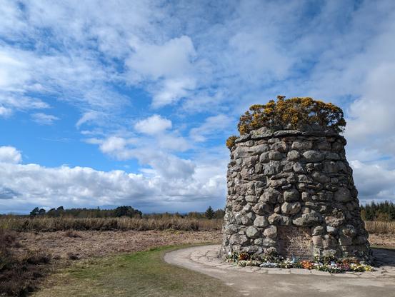 A stone cairn topped with yellow gorse stands to the right in a marshy field. Bouquets of flowers are laid around the base. The sky is blue and partly cloudy.