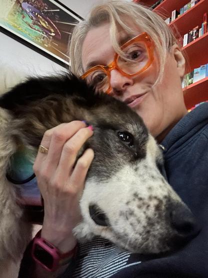 A large brown and white dog presses her head into the chest of a human with blond hair and bright orange glasses.