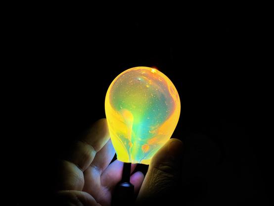 In the picture there is a colorful lightbulb, held by a hand, in front of a black background.