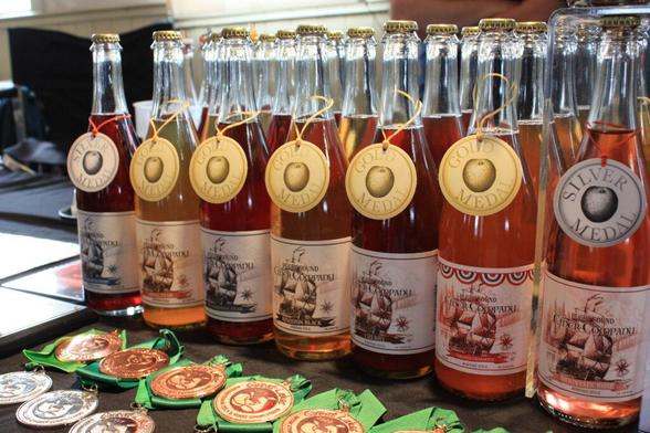 Bottles of various flavors of cider from the Port Townsend Cider Company, with gold or silver prize medallions hanging from or displayed in front of the bottles.