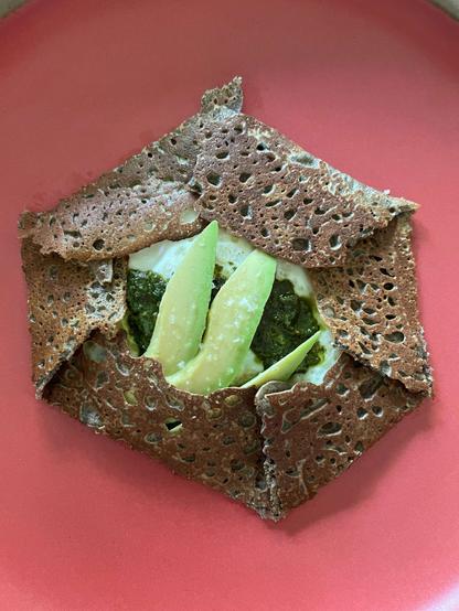 red plate with a neatly folded buckwheat crepe filled with avocado, pesto and a fried egg