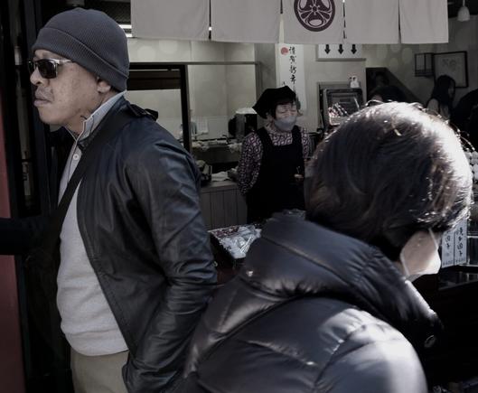 An old lady stands at the counter of a shop selling cakes in the street approaching Senso-ji Temple as people walk by.