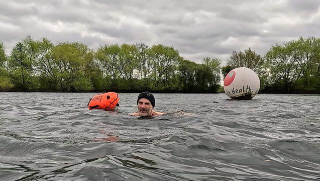 A scene in the middle of a lake. There are trees with fresh, green leaves around the edge at the back of the picture. There’s a man swimming - me! I’m wearing a black beanie and towing an orange float. You can just about see my shoulders and head, but the rest of me is underwater. My facial expression is a bit unreadable, but imagine the cold wind and slapping waves blowing from right to left in this picture, and you get the general idea. There’s a big, round white buoy behind me with the Whole Health logo of a red apple. After this, I had a nice long, hot sauna to warm back up. Bliss.