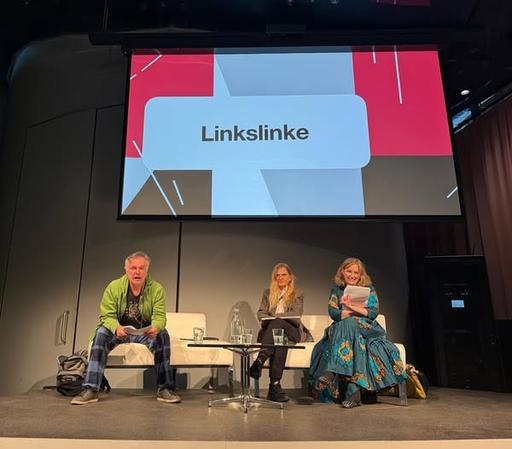 Die drei genannten Personen auf der Bühne, dahinter ist auf einer Videoleinwand "Linkslinke" zu lesen. Evelyn Schalk hat das Mikrophon.
