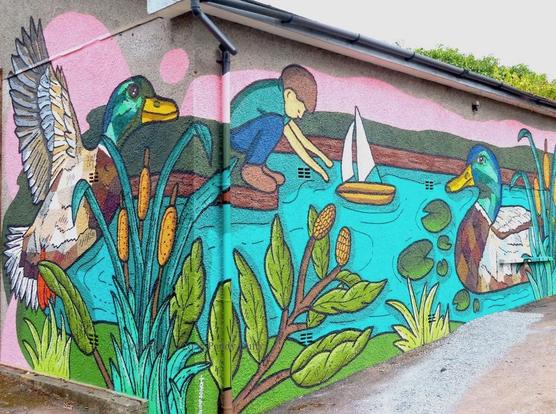 wall-painting on a corner wall showing the scene around a pond: (left) a duck takes flight; a child launches a toy boat (middle); a duck swims (right); a branch, lily-pads, and rushes complete the scene