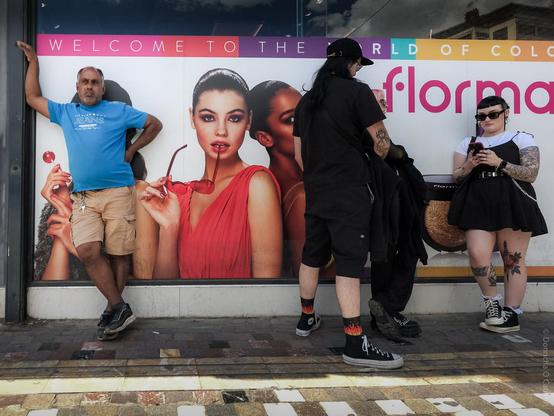 A candid street photo taken in Athens, Greece, showing four people standing in front of a colourful Flormar cosmetics advertisement. The ad features models with bold makeup and the text "WELCOME TO THE WORLD OF COLOURS flormar." On the left, a man in a blue t-shirt and beige shorts leans against the wall, while on the right, three people dressed in black, with tattoos and piercings, stand and interact, one looking at their phone.