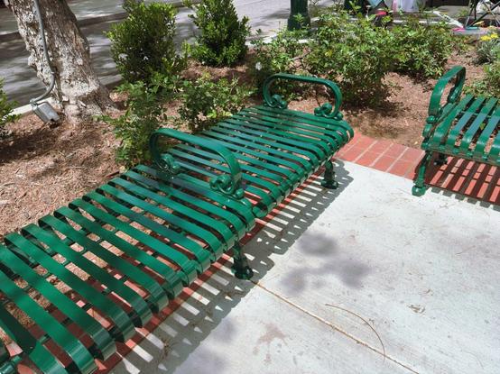A green metal park bench situated in a garden area. The bench has decorative scrollwork on the armrests and features horizontal slats forming the seat and backrest. It sits on a concrete and brick paved surface. Around the bench are small shrubs and plants growing in mulched soil. A tree trunk is visible on the left side with what appears to be a small light fixture attached to it. The bench casts distinct shadows on the concrete.