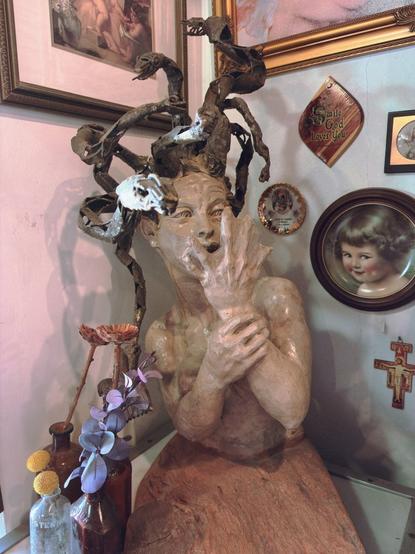 A sculptural bust on a reddish base. It depicts a Medusa-like figure with an open mouth and raised hands near its face in an expressive pose. The sculpture has metallic, snake-like structures extending from the head. It's displayed on a pink wall alongside various framed items including a decorative plaque reading "Smile God Loves You," a small crucifix, and a framed child's portrait. Small bottles and artificial flowers appear at the base of the display.