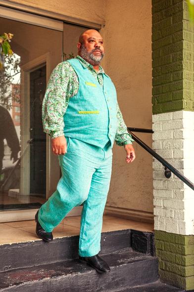 A bearish Black man descends the staircase in front of an apartment building. He has a stubbly shaved head and close cropped, salt & pepper beard. He wears perfectly fitted aqua linen trousers and vest with a floral paisely print dress shirt. The shirt’s print is in shades of green, blue, white, and gray and is complimented by the green and white painted bricks and charcoal painted steps of the building.