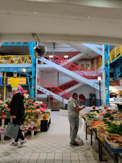 Puestos de frutas y flores en el mercado de Lehel. A la izquierda, uno de flores y a la derecha, uno de frutas. En cada puesto hay un hombre, mirando los productos. Al fondo se pueden ver unas escaleras con las barandillas metálicas de color rojo. Las barandillas de los pisos también son metálicas. La del primer piso amarilla y la del segundo, azul. Hay unas estructuras metálicas coloridas por todo el mercado.