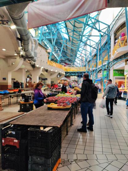 Vista general del mercado de Lehel. En primer plano hay un puesto con fresas y frutas. Una vendedora ofrece la mercancía a varios clientes. En el techo se aprecia la estructura metálica azul que se encuentra por todo el mercado.