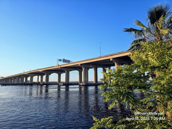 View across the Saint Johns River at a massive highway bridge spanning the wide river, beneath a big blue Florida sky reflecting onto the blue water below.