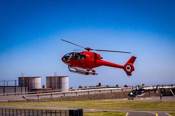 The image shows a red helicopter in flight above a landing pad. There is another helicopter on the ground in the background. The sky is clear, and there are some buildings or storage tanks visible.