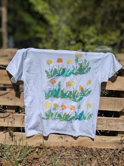 A big 5xl white gildan shirt with various shades of golden to orange dandelions printed on it in 3 rows, all dandelions in various stages of bloom and puff
