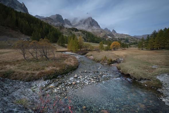 A mountainous landscape with a clear stream in the foreground and scattered trees with autumn foliage. Clouds partially obscure the peaks in the background.