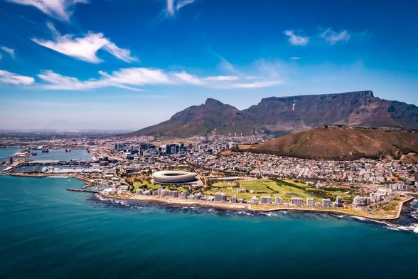 The image shows a scenic aerial view of Cape Town, South Africa. Visible landmarks include the Cape Town Stadium close to the shoreline and the iconic Table Mountain in the background. The image captures the city's coastline and urban landscape against a backdrop of mountains and a clear blue sky.