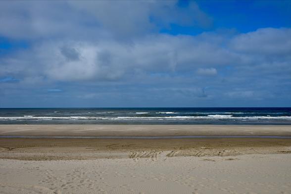 A wide view of a beach featuring sand, gently rolling waves, and a cloudy sky with varying shades of blue and gray.