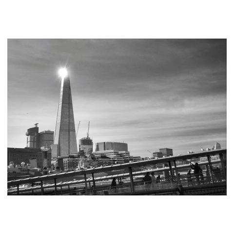This is a black and white landscape format image of The Shard in London. Taken from across the Thames, in the foreground are the roofs of jetties for boats to land, angled from around a quarter of the way up the frame on the right to the bottom left corner. This angle compliments the tall, thin pyramid shape of The Shard, which sits in the background around a third in from the left, towering above the surrounding buildings. Sunlight is catching and reflecting off the top of The Shard, creating a spotlight effect. Taken with a Sony RX100.