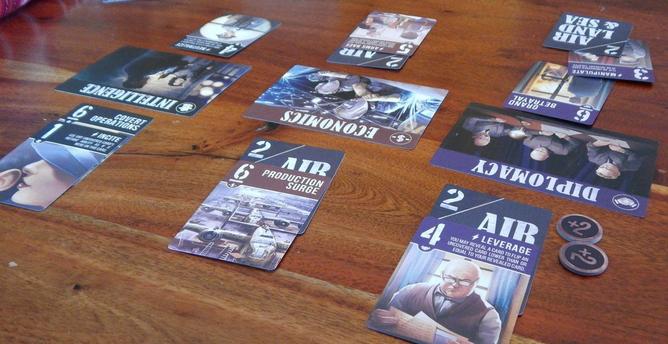 Reddish-brown wood tabletop with cards arrayed on it: three central “theater” cards with support cards on each side representing appropriate actions to gain power in each area. Round supply chits give bonuses in some areas.