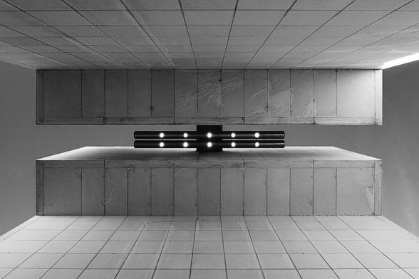 Symmetrical black-and-white photograph of a tiled ceiling with a modern light fixture made of two parallel black bars, each lined with five circular lights. The bars are mounted across a narrow vertical gap, through which the sky is visible, along with strips of sky on both the left and right sides, adding contrast to the structured interior.