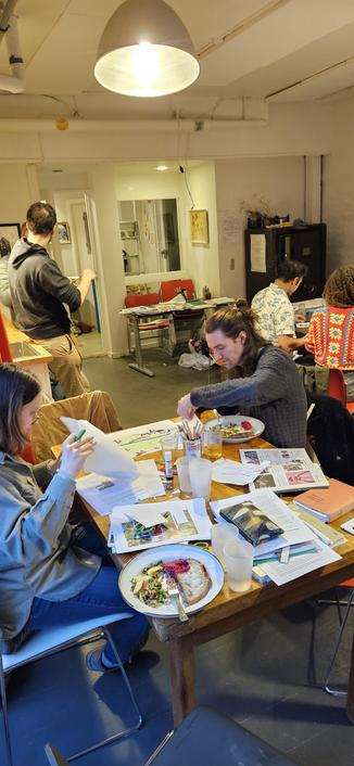 One person reads an article while another has a dinner plate and moves stuff on a collage. In the background people sit and talk and stand and talk.