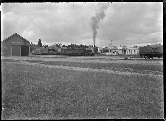 Black-and-white photo: Steam locomotive and train at Lumsden Railway Station. 1925. Description: Looking across at the train from the right-hand side. Across the road are a number of shops including “H. Casson, bootmaker”, “R. Martin, fruiterer & confectioner”, a chemist selling horse and cattle medicines, and “J. Jack, butcher”. Photographed by Albert Percy Godber. Citation: Godber, Albert Percy, 1875-1949: Collection of albums, prints and negatives. Ref: APG-1138-1/2-G. Alexander Turnbull Library, Wellington, New Zealand. https://natlib.govt.nz/records/22389195