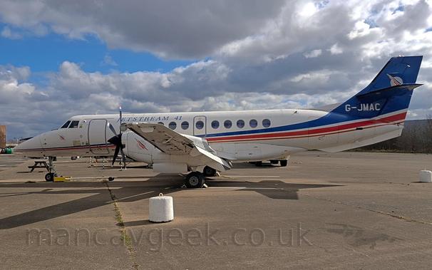 Side view of a white, twin propellor-engined airliner parked facing to the left on concrete.
There is a thin, blue, red, and orange stripe running along the body from the nose, getting wider as it moves rearwards, until it sweeps up towards the tail, which is almost entirely blue, with the registration "G-0JMAC" in white, and a white, winged, circular logo at the top.
There are dark blue "Jetstream 41" titles on the upper forward fuselage.
There is a white, concrete weight on the ground under the left wing-tip, that would normally be attached by a rope to the underside of the wing.
Grey concrete apron fills the bottom of the frame, , leading up to trees in the distance.
Fluffy white and grey clouds fill the top of the frame, with patches pf blue peeking through.