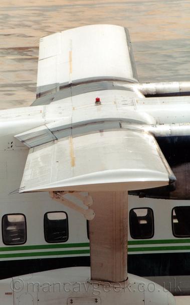 Close-up of the wing of a rather boxy, white, high-winged, twin propellor-engined airliner parked facing to the right.
There is a light and dark green stripe running along the body, under the almost-square passenger cabin windows.
The long, thin wings are supported by a white strut attached to a white pod attached to the bottom of the fuselage.
Grey concrete apron fills the background.