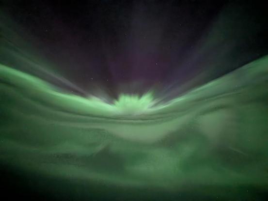 Foto de uma aurora, directamente acima de nós, dividindo o céu a meio - num lado uma névoa verde com o que parecem ondas, que parece explodir para o topo, em que vemos o céu escuro.