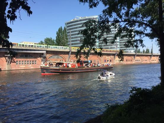 Eine malerische Aussicht mit einem Kanal mit einem großen Lastkahn namens "Andreas" und einem kleineren Boot. Im Hintergrund fährt ein Zug auf einem erhöhten Gleis neben modernen Gebäuden. Die Umgebung ist hell, mit klarem blauem Himmel und üppigem Grün.
