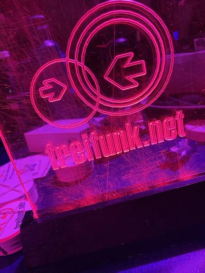 Ein rosa beleuchtetes Acrylschild mit dem Logo und der Webadresse "freifunk.net", das kreisförmige Designs und Pfeile enthält. Die Oberfläche zeigt Kratzer, und der Hintergrund ist dunk beleuchtet.