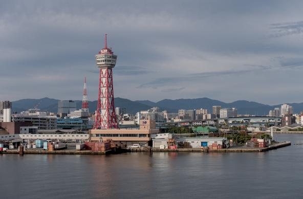 View from the sea of the Hakata port in Fukuoka City. The Hakata Port Tower, a red and white lattice tower resembling the Tokyo Tower, stands in the middle of the picture. The surface of the sea is quiet and reflects the color of the port authority's buildings and palm trees.
A freeway links the port to the city behind it, and its background is a landscape of forested hills.
The sky is cloudy but the morning sunlight is still quite bright.